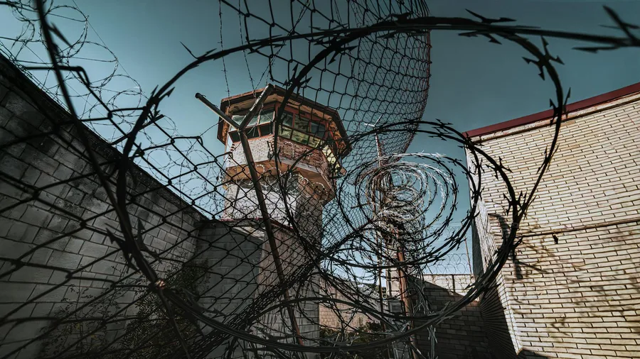 A dramatic view of a prison watchtower seen through coiled barbed wire under a clear sky.