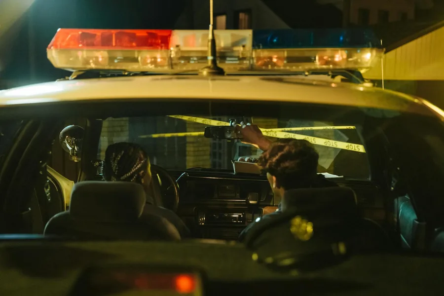 Police officers monitoring a scene through rearview mirror at night with caution tape visible.