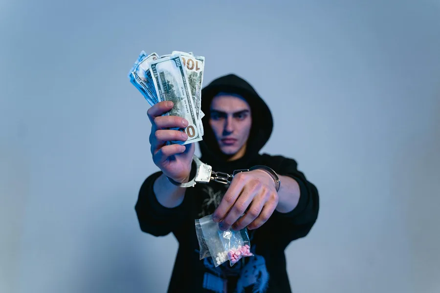 Handcuffed man in a hoodie holding cash and drugs, depicting crime and addiction.