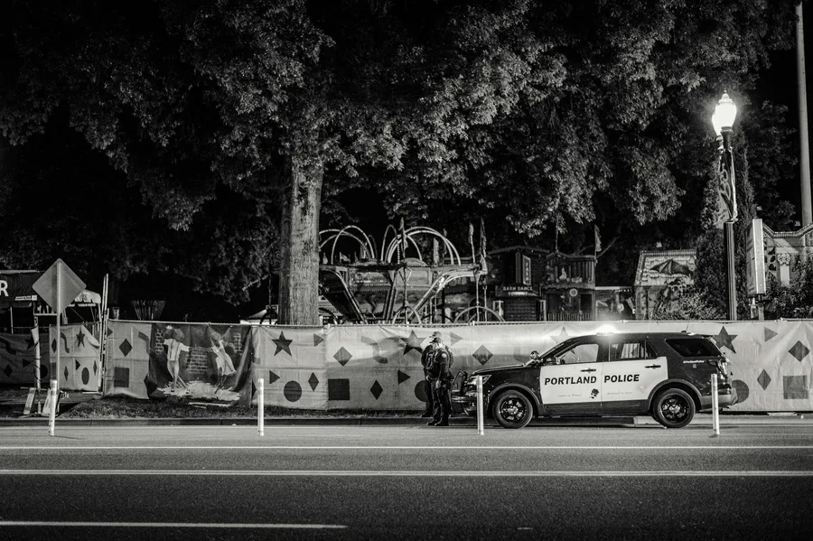 Black and white nighttime street view featuring a Portland police car parked by a decorated fence.