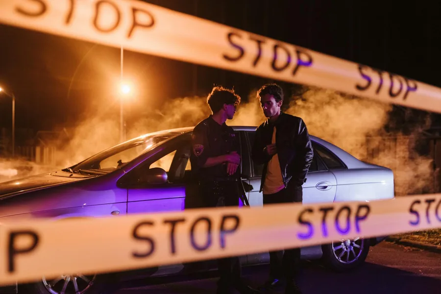 Nighttime crime scene with stop tape and two men by a car surrounded by smoke.