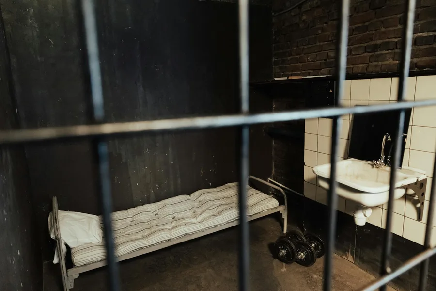 Moody prison cell interior featuring a metal bed, sink, and dumbbells behind bars.