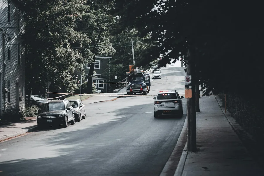 Police vehicle driving on a tranquil city street, surrounded by trees and parked cars.