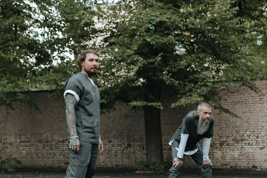 Two men standing outdoors in a prison yard with a brick wall and trees.