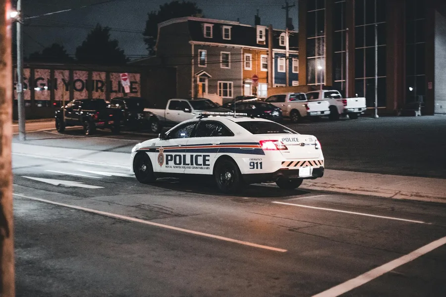 A police car drives through an urban street at night illuminated by streetlights.