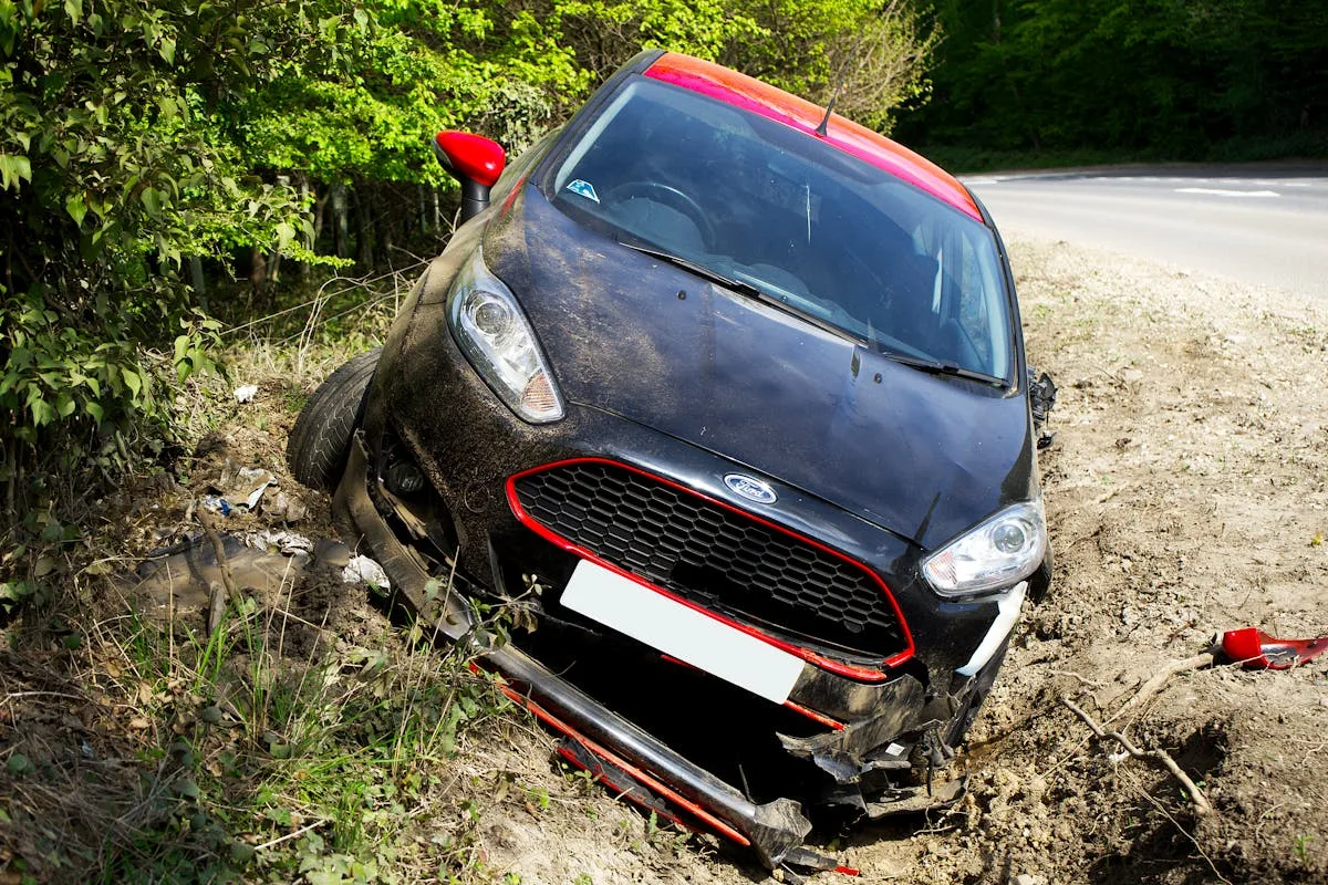 A black car, seemingly a Ford, crashed and damaged in a roadside ditch with surrounding greenery.
