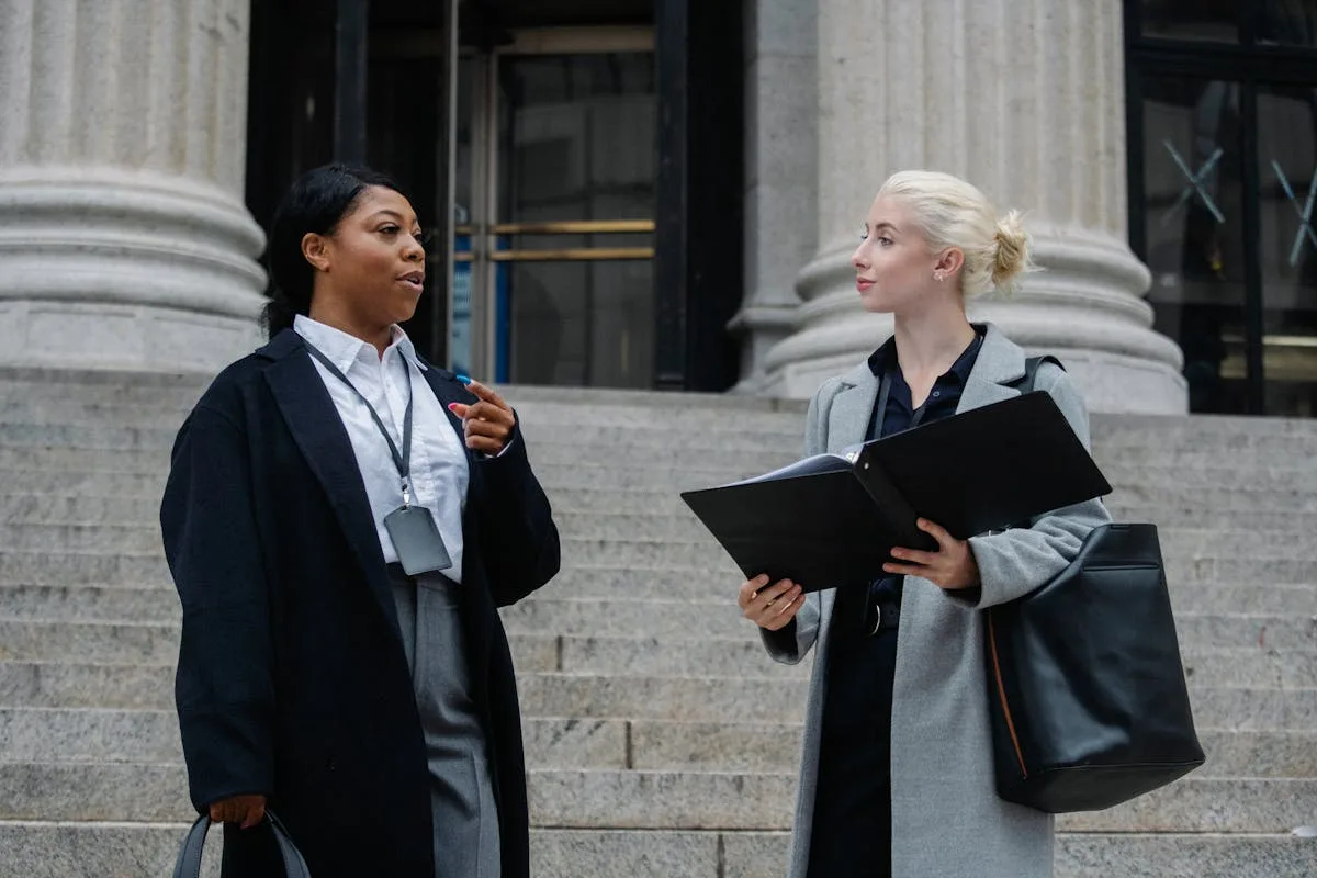 Two professional women in discussion on outdoor courthouse steps, holding documents.