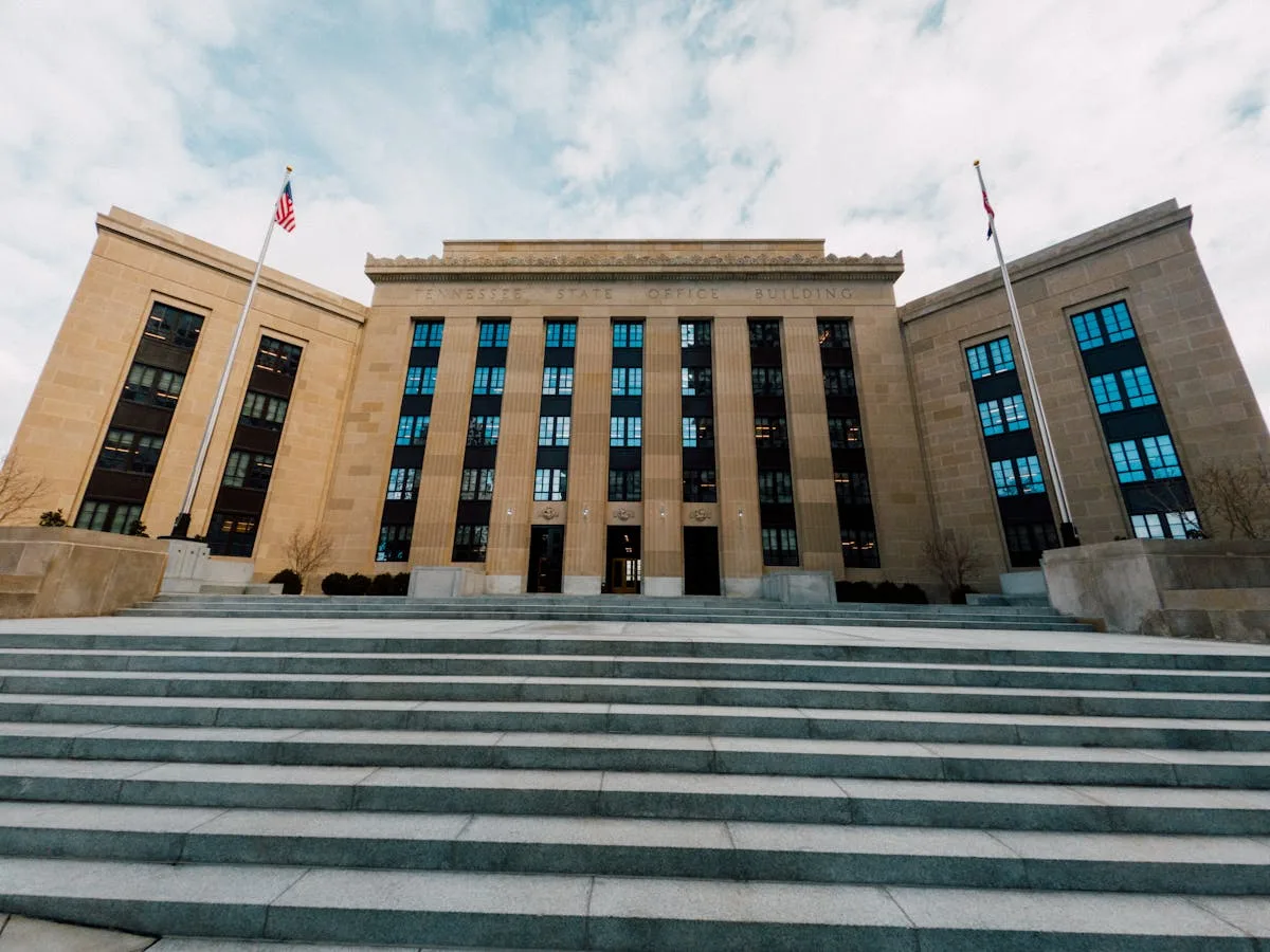 Federal style government building with flags and stairway under cloudy sky.
