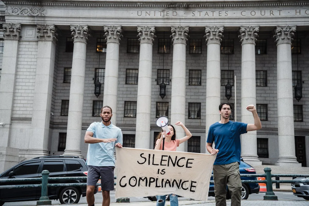Three protesters with a 'Silence is Compliance' banner in front of a courthouse.
