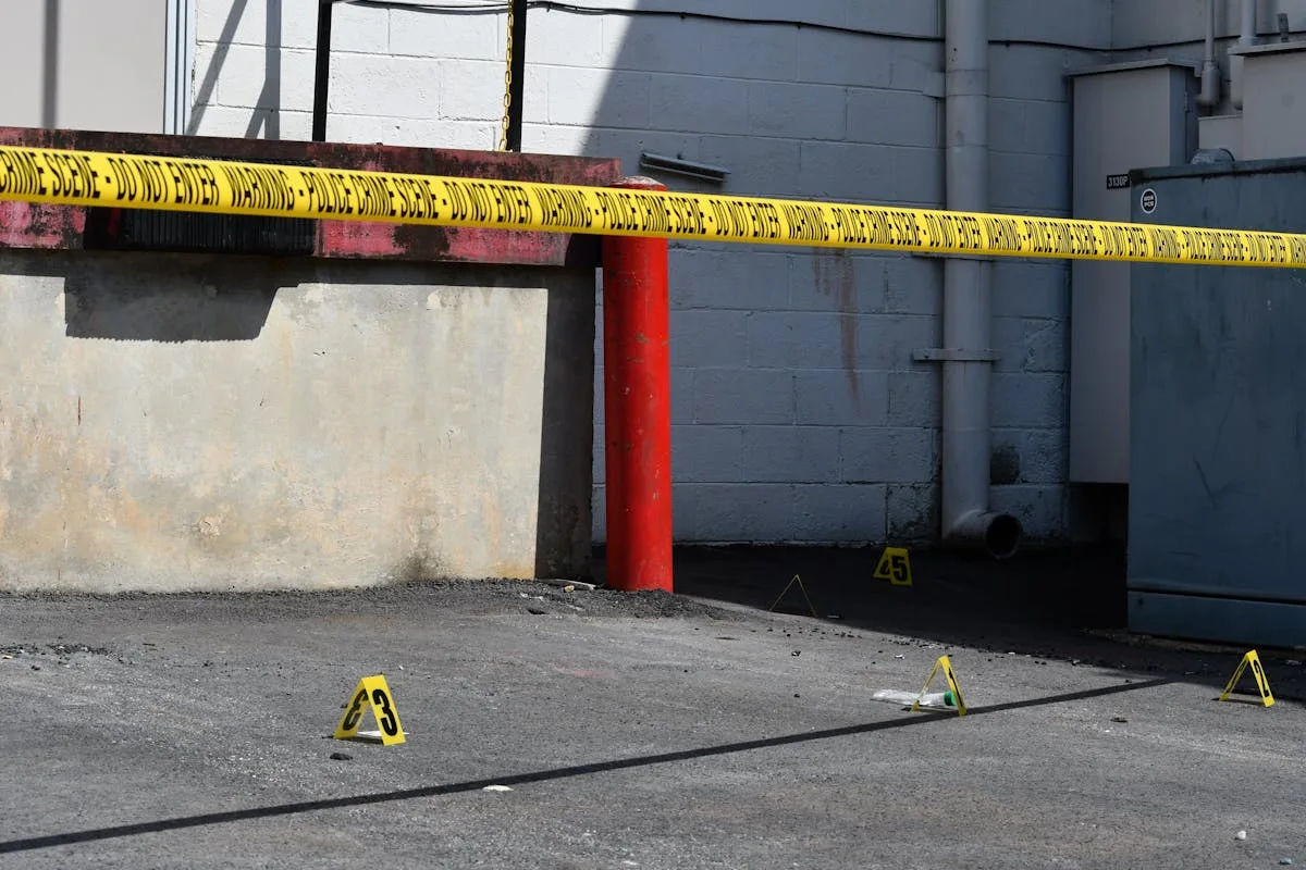 Sunlit urban scene showing a crime scene with caution tape and evidence markers, conveying investigation atmosphere.
