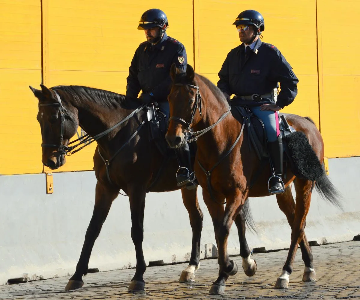 Two mounted police officers riding horses beside a yellow building wall.
