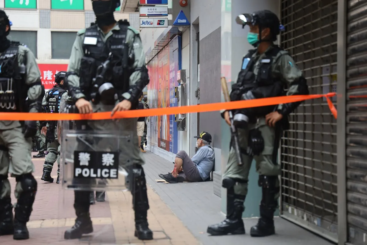 Urban environment with police presence and a civilian sitting against a wall, showing tension.