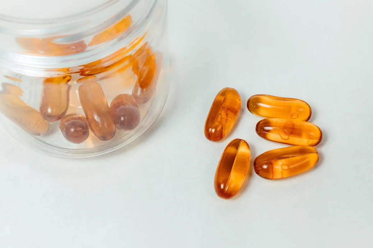 Glass container with amber capsules spilling out on a white background.
