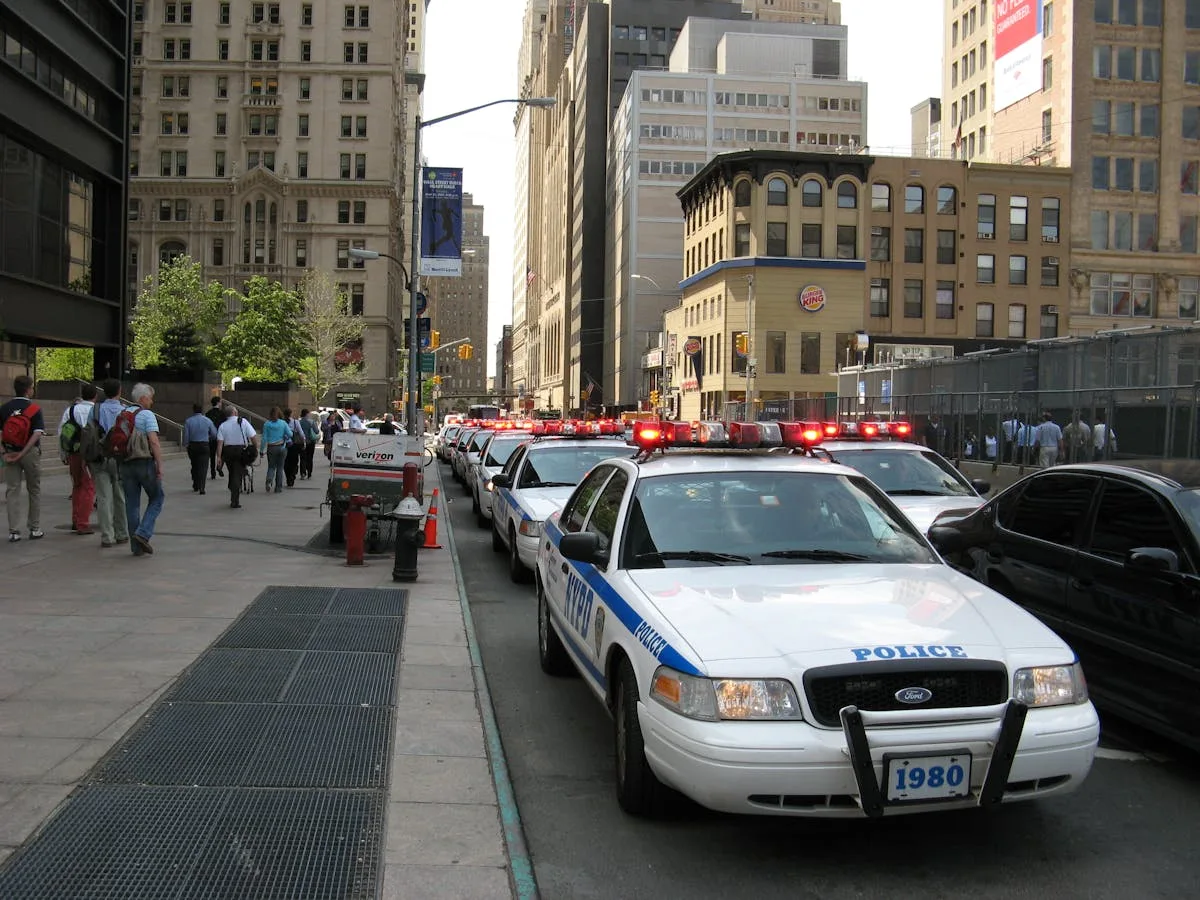 A line of NYPD police cars with flashing lights on a busy New York street, featuring pedestrians and tall buildings.
