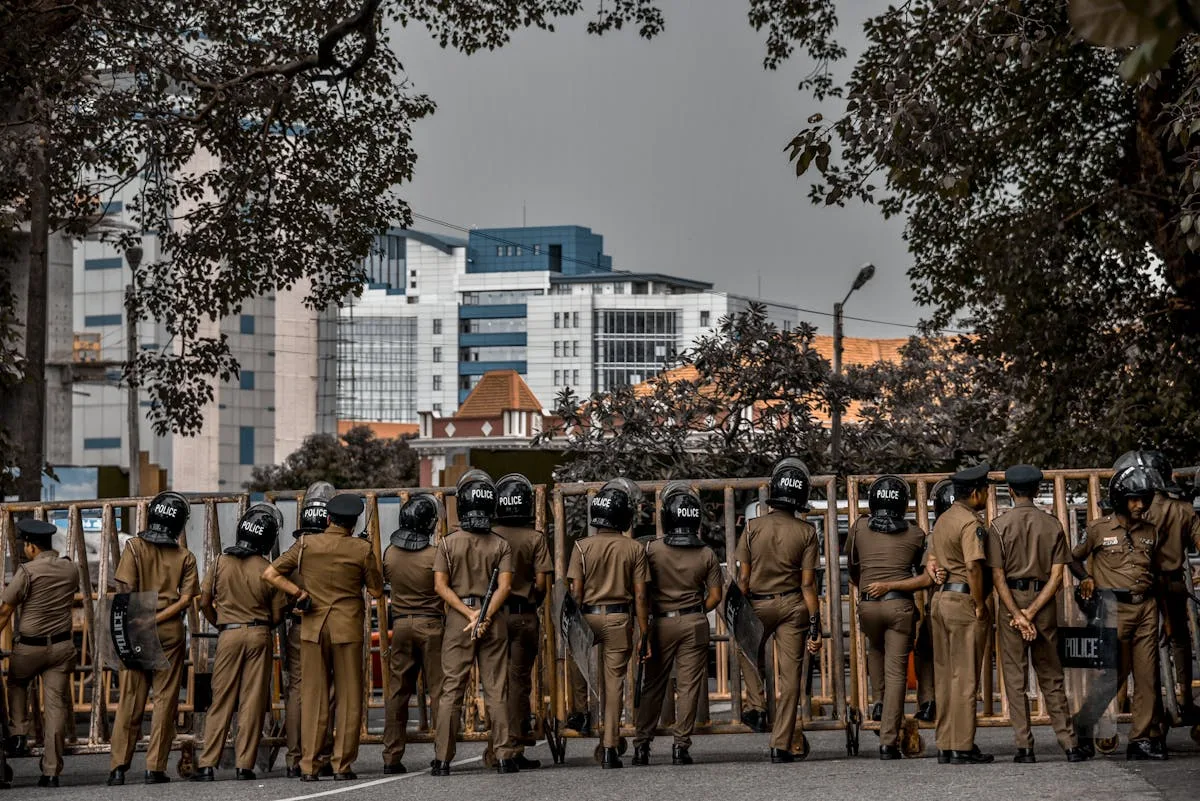Back view of police officers in uniform standing on a city street surrounded by buildings and trees.