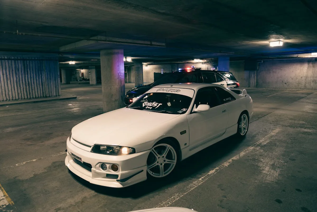 A white sporty car parked in an underground garage, nearby police car with light on.
