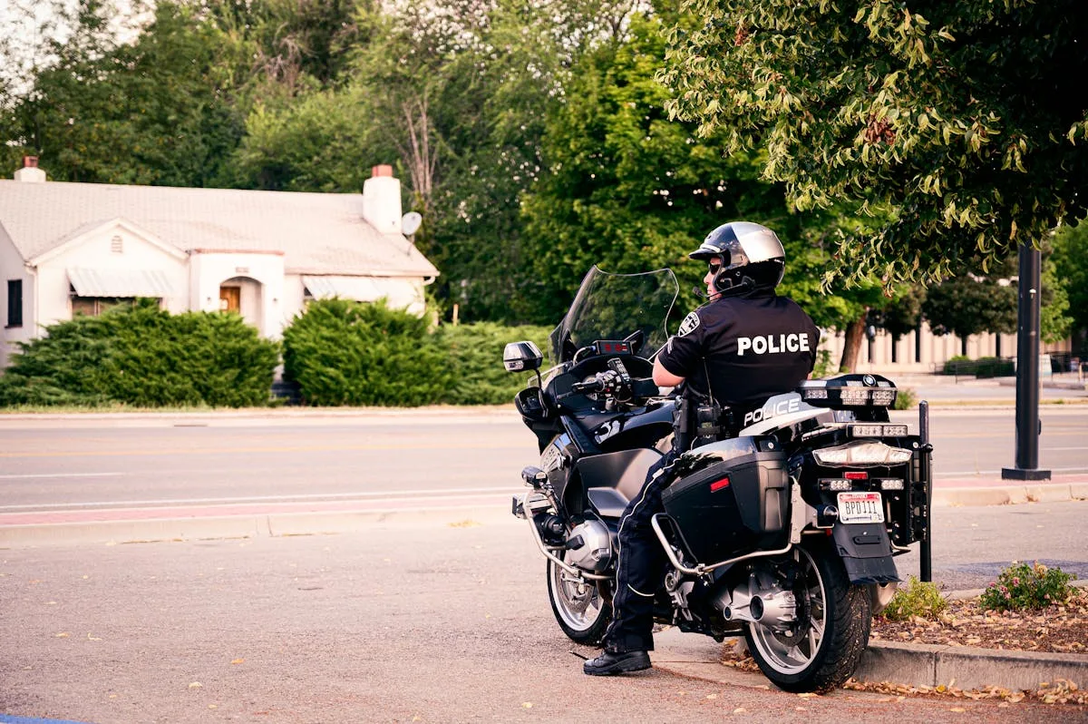 A police officer on a motorbike observes a quiet suburban street lined with greenery.
