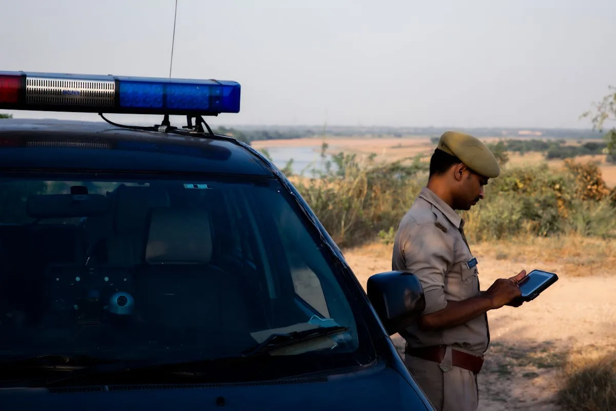 A police officer using a tablet beside a patrol car in a rural outdoor setting.

