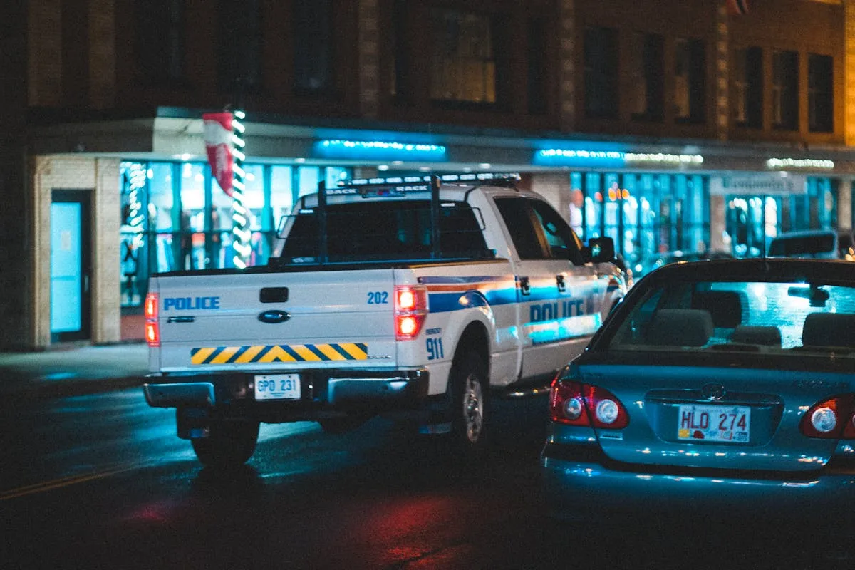 Police pickup with tail light riding on asphalt roadway near car along building with glass showcase in city on evening time
