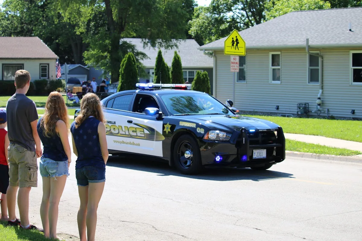 A police car on patrol in a suburban street with onlookers watching nearby.