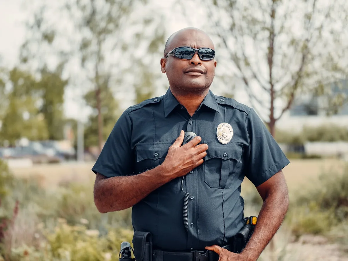 Confident police officer in uniform outdoors, hand on badge, sunny park setting.
