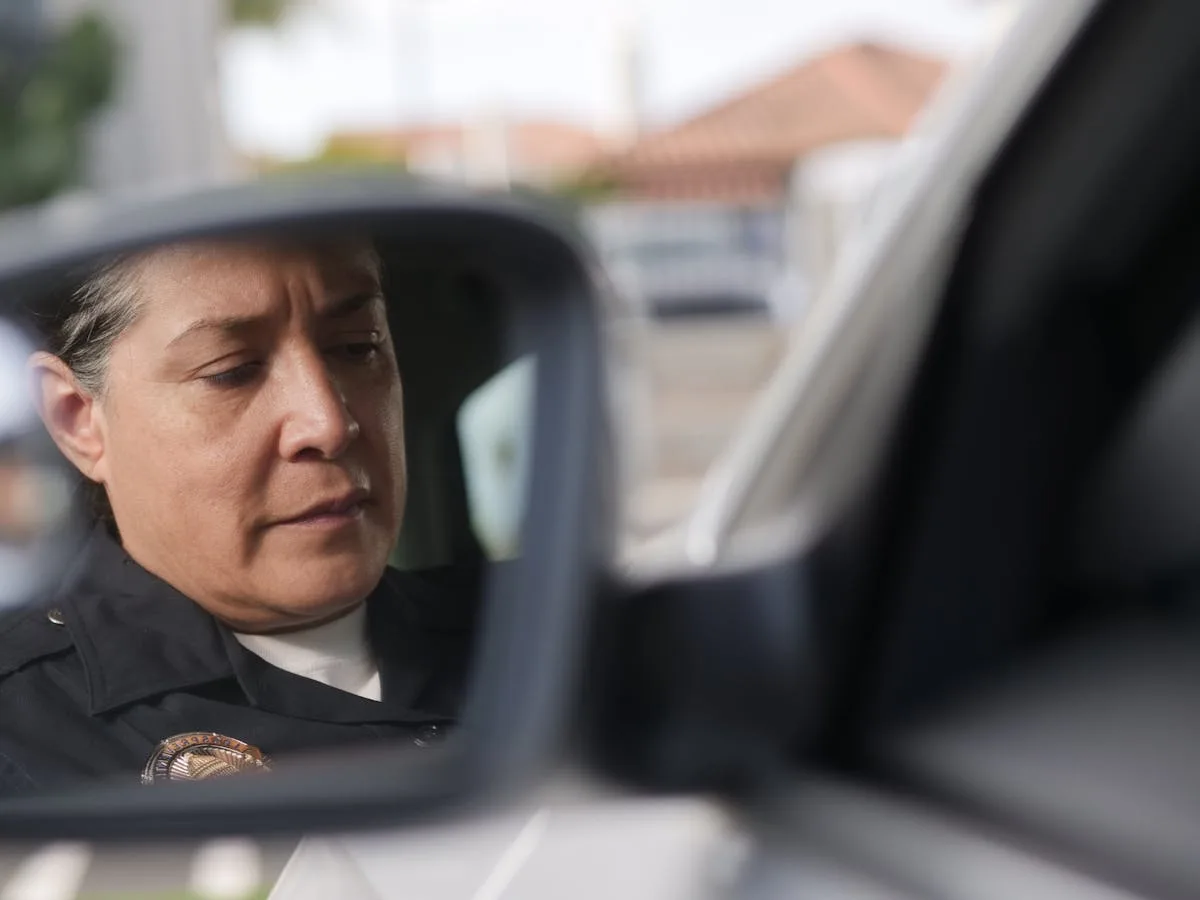 A police officer focused while appearing in a car mirror reflection.