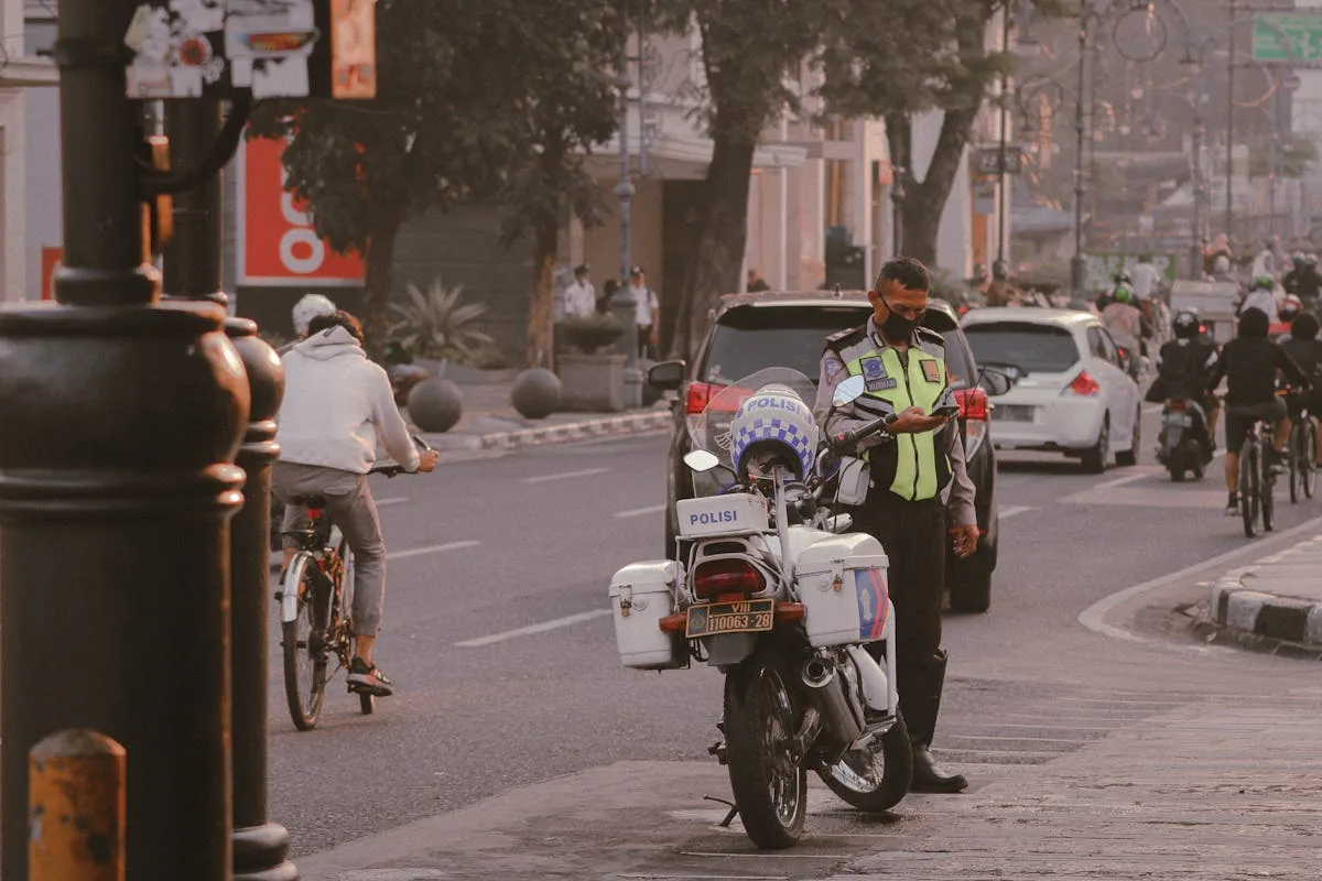 A police officer checks his phone near motorcycles on a busy city street with cyclists and cars.
