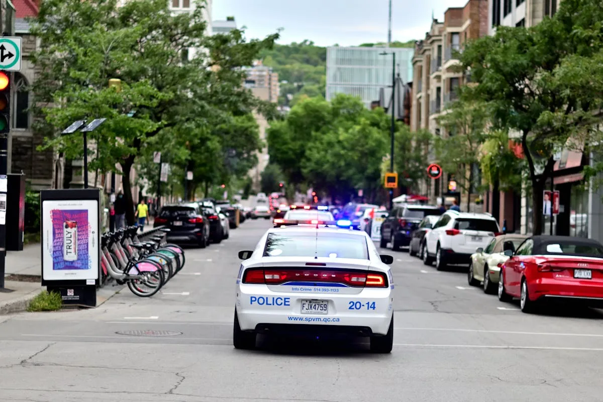 Police car driving through urban street lined with trees and parked bicycles.
